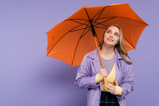 dreamy young woman in trench coat standing under orange umbrella on purple  - Фото, изображение