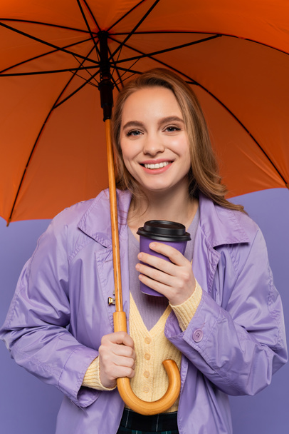 happy young woman in trench coat holding paper cup and standing under orange umbrella isolated on purple  - Valokuva, kuva
