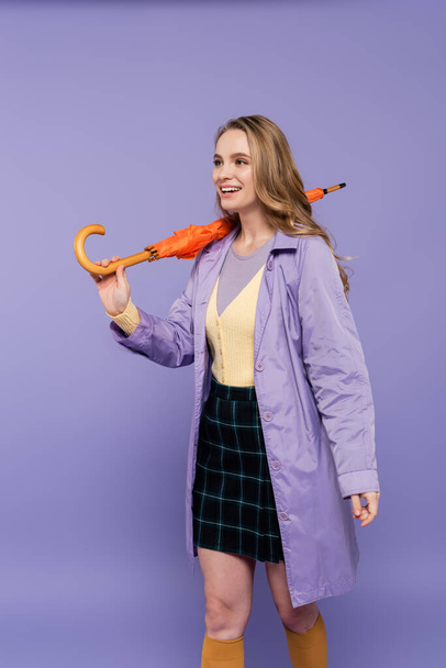 positive young woman in trench coat standing with orange umbrella isolated on purple  - Foto, afbeelding