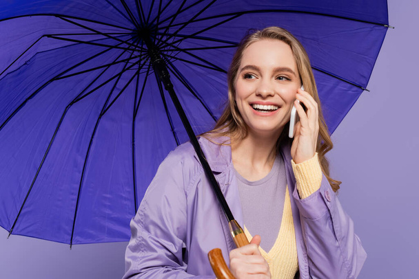 happy young woman in trench coat standing with blue umbrella and talking on smartphone isolated on purple  - Photo, Image