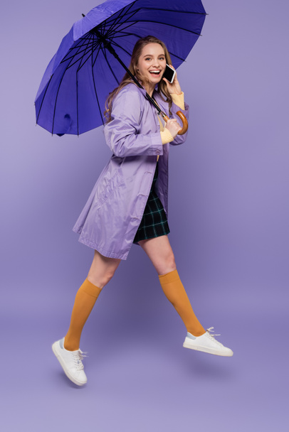 Trençkotlu, mavi şemsiyeli ve mor üzerine akıllı telefondan konuşan mutlu genç bir kadın.  - Fotoğraf, Görsel