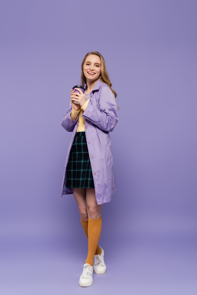 full length of happy young woman in trench coat holding paper cup on purple  - Foto, Bild