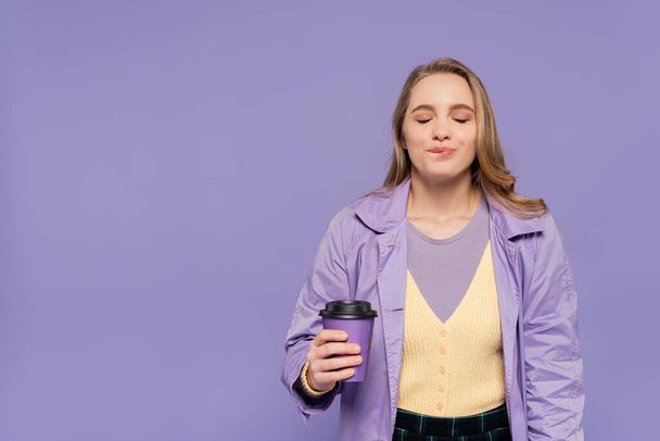 pleased young woman in trench coat holding paper cup and biting lip isolated on purple  - Photo, Image