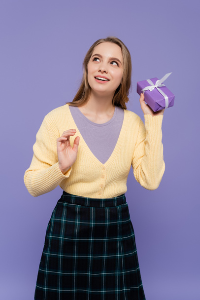 dreamy young woman holding wrapped gift box isolated on purple - Фото, зображення