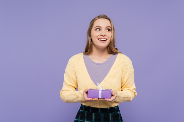 збуджена молода жінка тримає загорнуту подарункову коробку ізольовано на фіолетовому
 - Фото, зображення