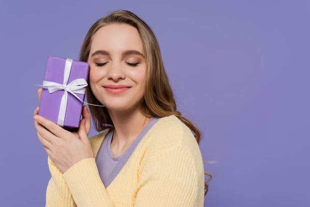 pleased young woman holding wrapped gift box isolated on purple - Фото, изображение