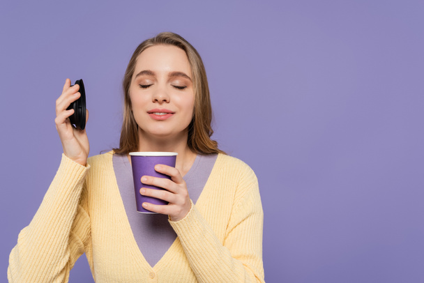 joven mujer oliendo café para ir en taza de papel aislado en púrpura - Foto, imagen