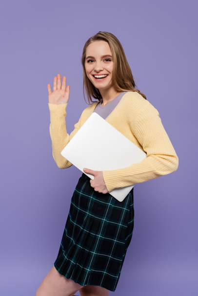 χαρούμενο μαθητή κουνώντας το χέρι και κρατώντας φορητό υπολογιστή απομονωμένο σε μωβ  - Φωτογραφία, εικόνα