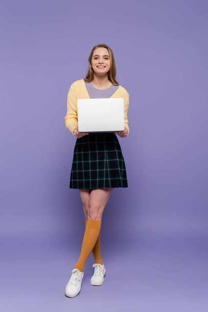 full length of cheerful young woman holding laptop on purple  - Foto, Imagen