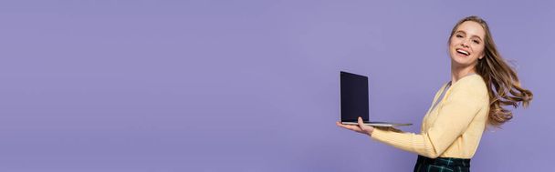 веселая молодая женщина держит ноутбук с чистым экраном изолированы на фиолетовый, баннер - Фото, изображение