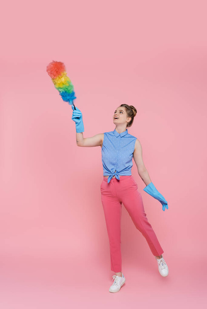 pełna długość wytatuowanej młodej kobiety w niebieskich gumowych rękawiczkach trzymającej szczotkę do kurzu na różowym  - Zdjęcie, obraz