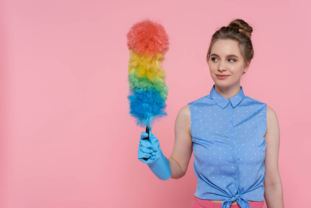smiling young woman in blue rubber glove holding dust brush isolated on pink  - Photo, Image