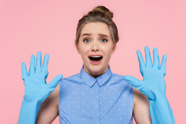wstrząśnięta kobieta pokazuje ręce w niebieskich gumowych rękawiczkach odizolowanych na różowo  - Zdjęcie, obraz