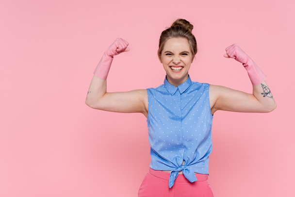 счастливая и татуированная женщина в резиновых перчатках, показывающая силу, изолированную на розовом  - Фото, изображение