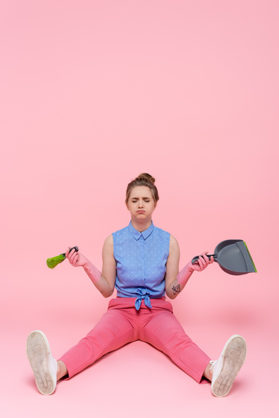 full length of sad young woman in rubber gloves holding broom and dustpan while sitting on pink - Фото, изображение