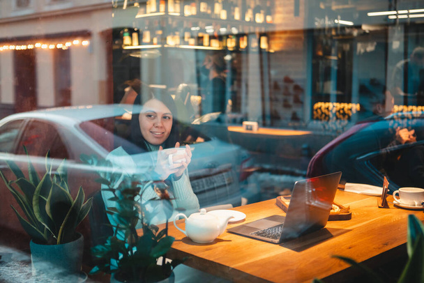 Restoranda dizüstü bilgisayarda çalışan gülümseyen bir kadın çay içip bardaktan hamburger yiyor. - Fotoğraf, Görsel