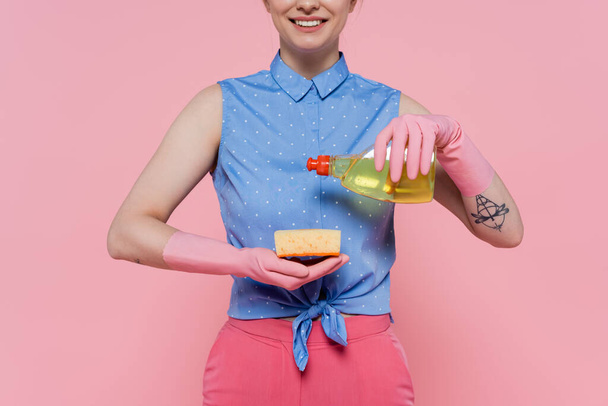 osittainen näkymä onnellinen ja tatuoitu nainen kaatamalla pesuainetta sienellä eristetty vaaleanpunainen  - Valokuva, kuva