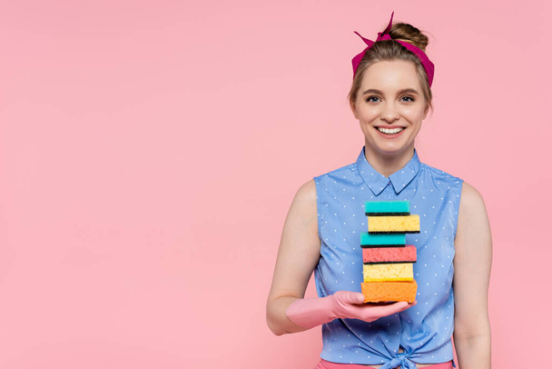 happy young woman in rubber gloves holding stacked sponges isolated on pink  - Фото, зображення