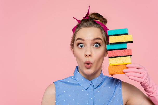 surprised young woman in rubber glove holding stacked sponges isolated on pink  - Photo, Image