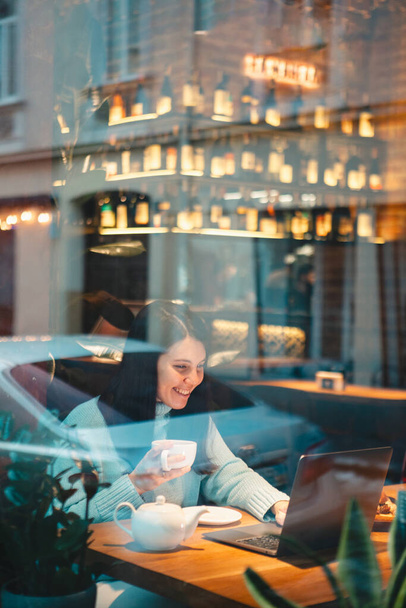 donna sorridente che lavora sul computer portatile nel ristorante bere tè mangiare hamburger vista attraverso il vetro - Foto, immagini