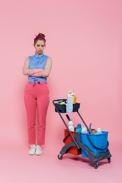 full length of offended young woman in rubber gloves standing with crossed arms near housekeeping cart on pink - Foto, immagini