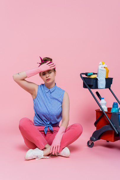 piena lunghezza della giovane donna stanca in guanti di gomma seduta vicino al carrello delle pulizie con prodotti per la pulizia su rosa - Foto, immagini