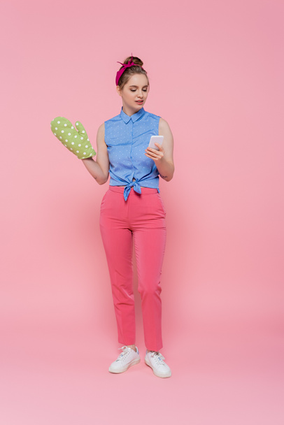 full length of young woman with green baking glove holding smartphone isolated on pink - Фото, изображение