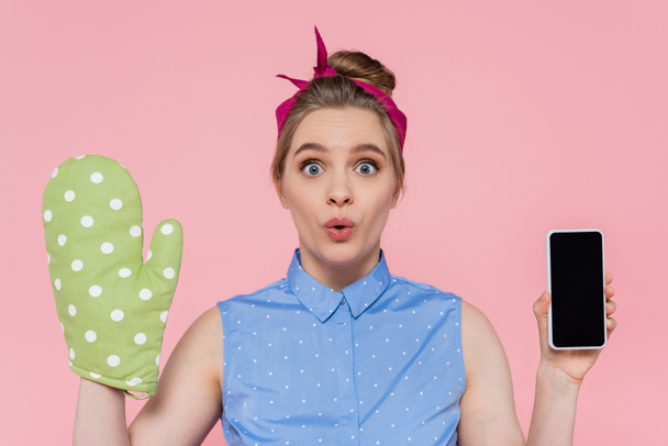 surprised young woman with green baking glove holding smartphone with blank screen isolated on pink - Photo, image