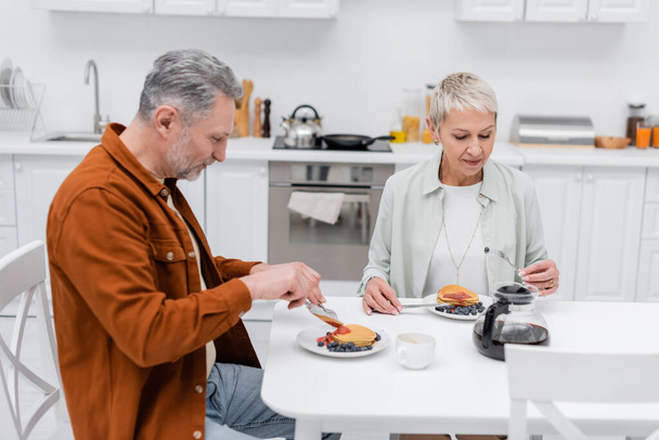 Mature man cutting pancakes near wife and coffee in kitchen  - Valokuva, kuva