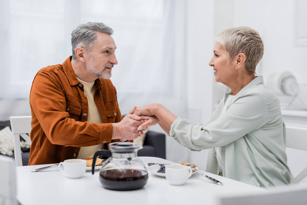Positive couple holding hands near coffee and pancakes in kitchen  - Fotoğraf, Görsel