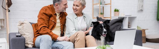 Smiling couple talking and petting border collie near laptop at home, banner  - Photo, Image