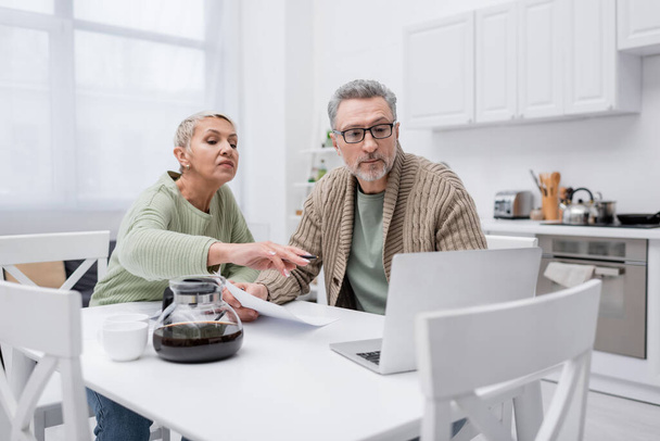 Elderly woman pointing at laptop near husband with paper and coffee in kitchen  - Фото, изображение