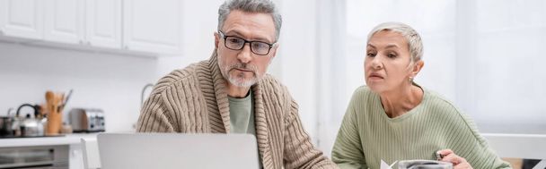 Mature man looking at blurred laptop near wife in kitchen, banner  - Фото, зображення