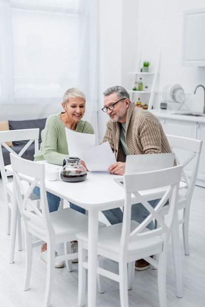 Cheerful senior woman holding paper near husband, laptop and coffee in kitchen  - Фото, изображение