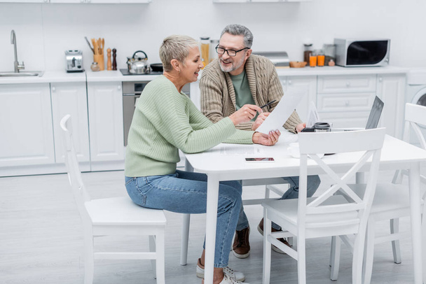 Positive pensioners holding documents near devices in kitchen  - Φωτογραφία, εικόνα