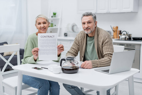 Pensioners holding contract and looking at camera near devices and coffee in kitchen  - Photo, image