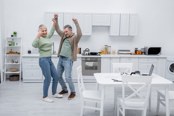 Happy couple dancing near papers and gadgets in kitchen  - Фото, изображение