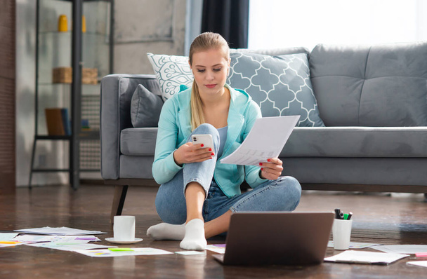 Молодая женщина работает с документами, используя ноутбук, сидя дома на полу. Студентка, предприниматель или девушка-фрилансер, работающая или обучающаяся дистанционно через интернет. - Фото, изображение