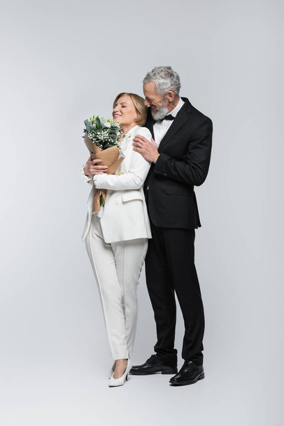Mature groom hugging cheerful bride with bouquet on grey background  - Fotografie, Obrázek