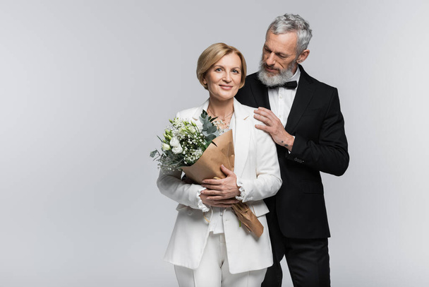 Mature groom in suit hugging smiling bride with wedding bouquet isolated on grey  - Fotó, kép