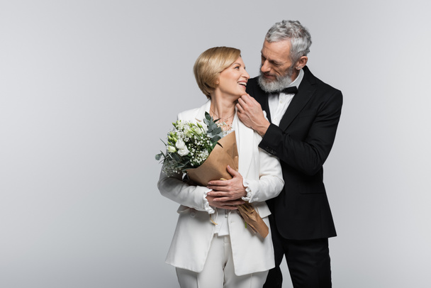 Mature groom touching chin of smiling bride with bouquet isolated on grey  - Fotó, kép