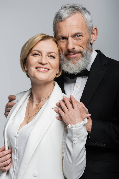 Portrait of middle aged groom hugging smiling bride and looking at camera isolated on grey  - Фото, зображення