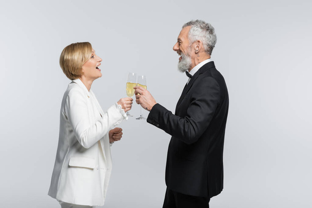 Vista laterale della sposa positiva clinking champagne con lo sposo maturo isolato su grigio  - Foto, immagini