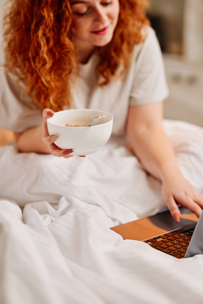 A redhead girl eating breakfast in the bed. A ginger girl sitting in her bed in the bedroom and eating cereals while surfing on internet on a laptop. - Photo, Image