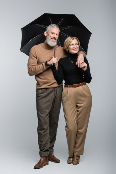 Stylish mature man holding umbrella and hugging cheerful wife on grey background  - Foto, imagen