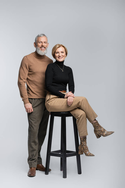 Homme élégant souriant regardant la caméra près de la femme assise sur une chaise sur fond gris  - Photo, image