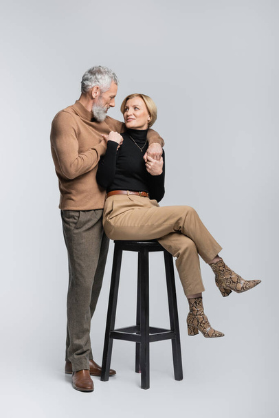 Reifer Mann umarmt stilvolle Frau sitzt auf Stuhl auf grauem Hintergrund  - Foto, Bild
