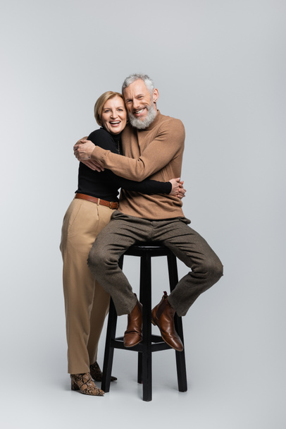 Happy and trendy mature couple hugging and looking at camera on grey background  - Valokuva, kuva