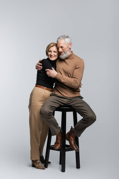 Smiling mature woman hugging husband sitting on chair on grey background  - Zdjęcie, obraz