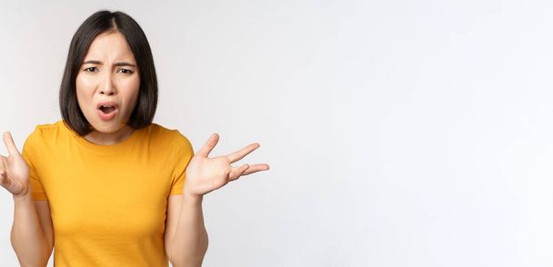 Frustrated, confused asian woman cant understand smth, looking clueless, shrugging shoulders, standing in yellow tshirt over white background - Foto, Bild